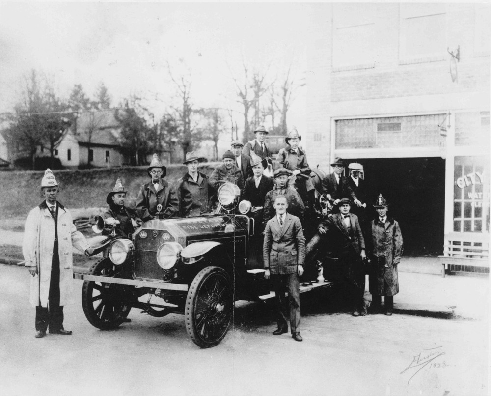 About fifteen firemen in and around a firetruck in front of a firehouse with the garage door open, circa 1913-1930.