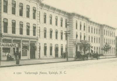 Un largo edificio blanco de tres pisos al lado de un restaurante. Delante hay un carruaje. El texto de la imagen dice "Yarborough House Raleigh, N.C."