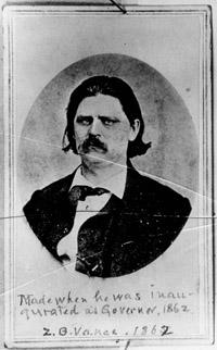 Zebulon Vance, a white man with chin length hair and a mustache. He is wearing a dark coat, white collared, button-up shirt and dark neckcloth. He has a serious expression.