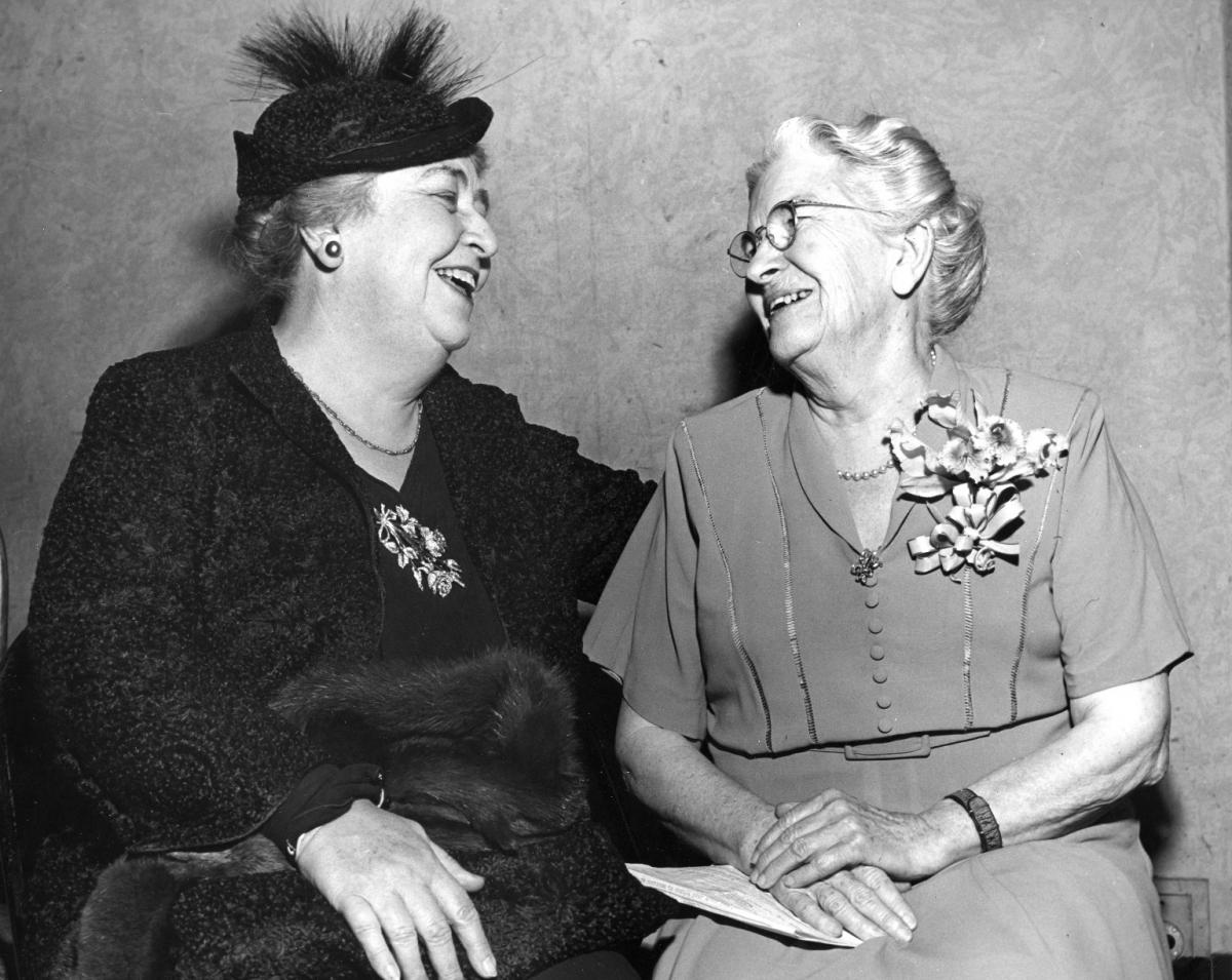Two women sitting and smiling at each other.  They are older. The woman on the left is dressed in a dark dress and hat. The woman on the right is older with gray hair and glasses.  She is wearing a light colored dress and has flowers pinned on the left side of the dress top.