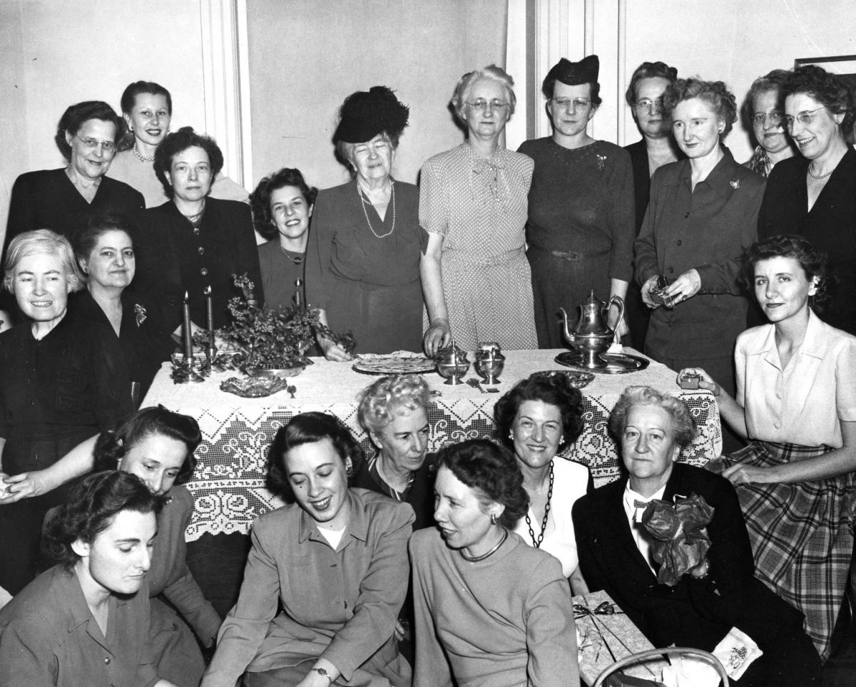 A group of women pose around a lace covered dining table. The table is set with a silver tea service, a plate of thin wafers, and a Christmas decoration. The women are all dressed up and some hold Christmas presents. Jane McKimmon is standing in the center back. She has white hair and glasses and wearing a short-sleeved polka-dotted dress. Ruth Current is sitting to the front of the table directly in front of McKimmon. She has dark hair and long chain-link necklace.