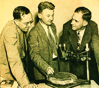 Three men standing next to a microscope with a Dare stone underneath.