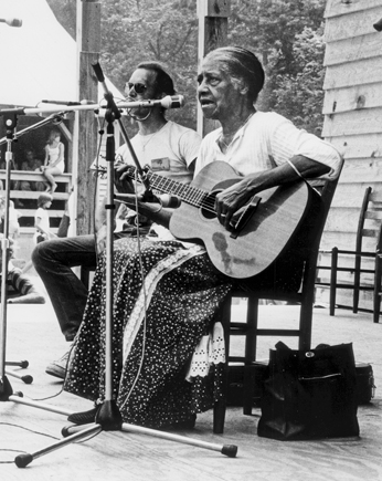 Elizabeth Cotten sings into a microphone.  She is wearing a long skirt and white shirt.  She is strumming a guitar.  Her hair is pulled back to the nape of her neck. She is older.