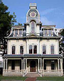 A three-story house with a square, Mansford style roof. A low, square dome is in the center of the roof. A covered porch extends along the first floor. Steps lead up to a wood door.