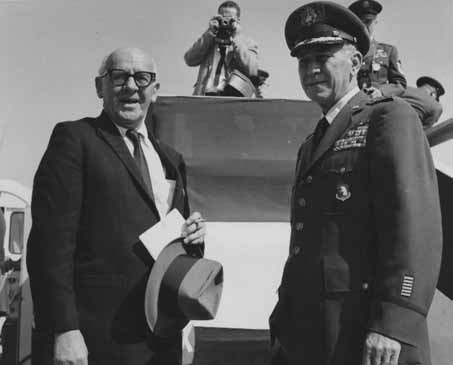  General Frank A. Armstrong, Jr. and an unidentified man standing together. Armstrong is in an air force uniform coat, dress, shirt, tie, and hat. Photographer and other military officers behind them.
