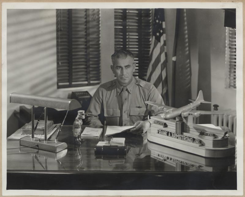 General Frank Armstrong seated at desk in his office. A model plane sits on the desk, a U.S. flag is in the background.