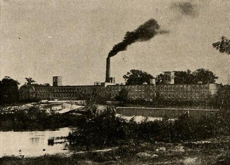 Rocky Mount Mills. A large factory complex with an active smokestack is in the background and small shrubs dot the foreground.