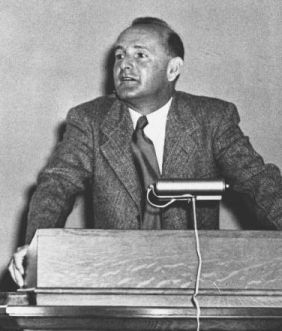 Black and white photograph of a white man with short hair wearing a suit and tie. He is standing at a lectern with a lamp on it and appears to be speaking to an audience.