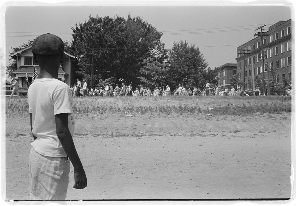 Photograph shows a young black boy watching a group of people, some carrying American flags, march past to protest the admission of the "Little Rock Nine" to Central High School.