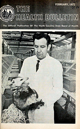 The cover of The Health Bulletin, February 1971. Dr. Jurgelsky removes an opossum from a nest box in the building developed at the National Institute of Environmental Health Sciences, Research Triangle Park, to house the opossum breeding colony. Individual cages and nest boxes are seen to either side. In order to avoid a painful, and often severe bite on the hand, the animals must be restrained by quickly and firmly grasping the nape of the neck. Magazine cover depicting a man in a white coat holding an opossum by the nape of its neck.