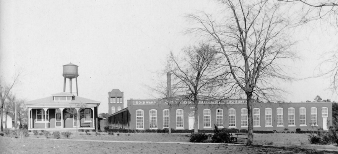 Black and white photo of the Clayton Cotton Mill, showing a small brick building to the left with a water tower, and a larger brick building to the right.