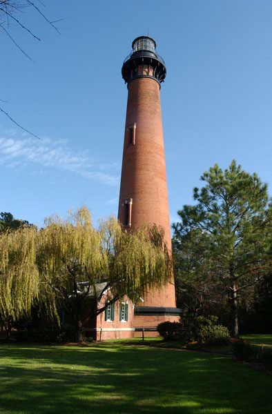 Photograph of the Currituck Beach Lighthouse, a brick lighthouse, against a clear blue sky surrounded by pine and other trees.