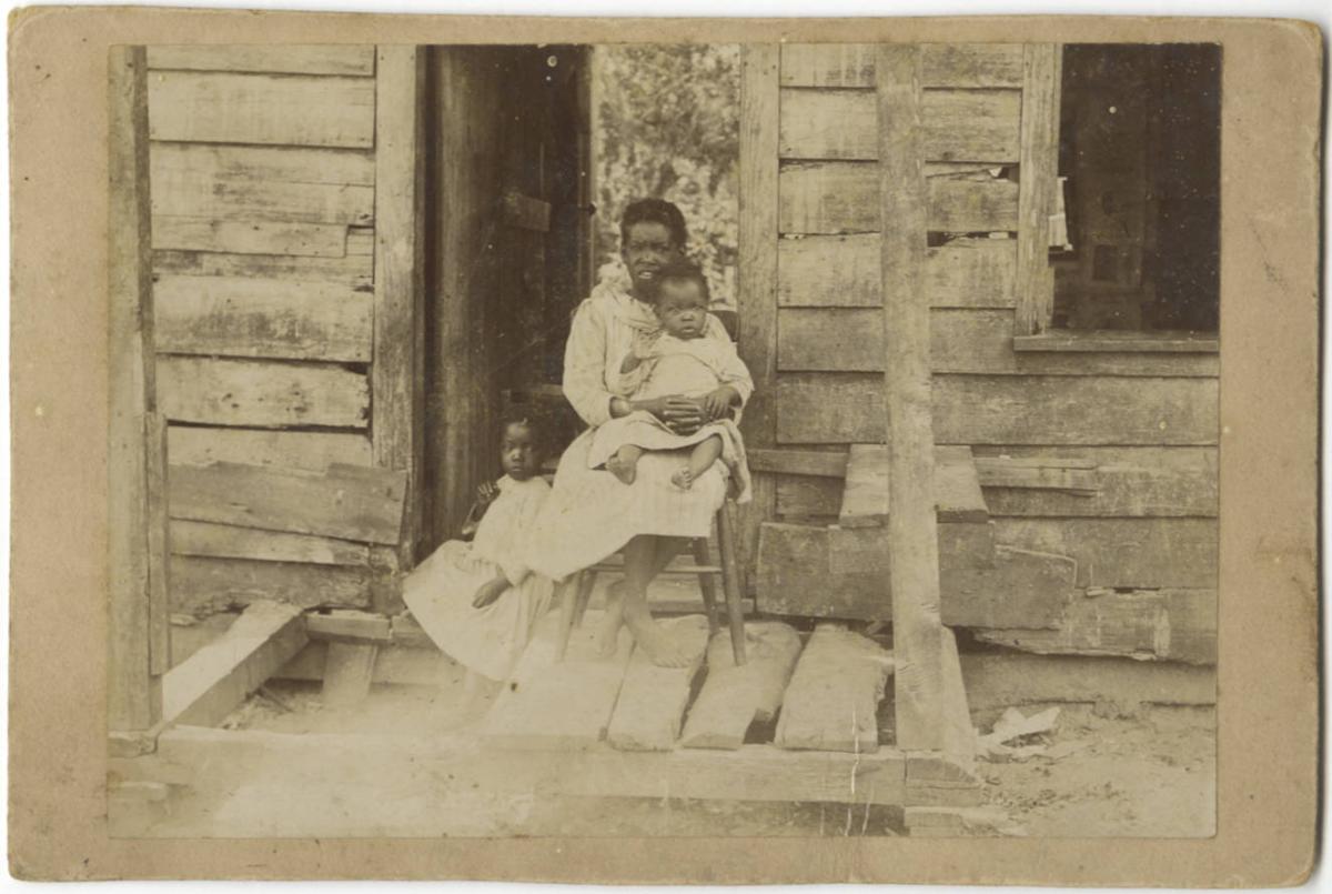 Three children on a porch in front of a wooden-sided home. All three children are black with short hair and white dresses. The oldest child is seated in a chat, holding a toddler in her lap, while another young child sits beside on the porch.