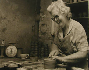 Una mujer creando un cuenco de cerámica. Lleva delantal y gafas. Ella está sonriendo, su cabello es blanco. Ella es mayor.