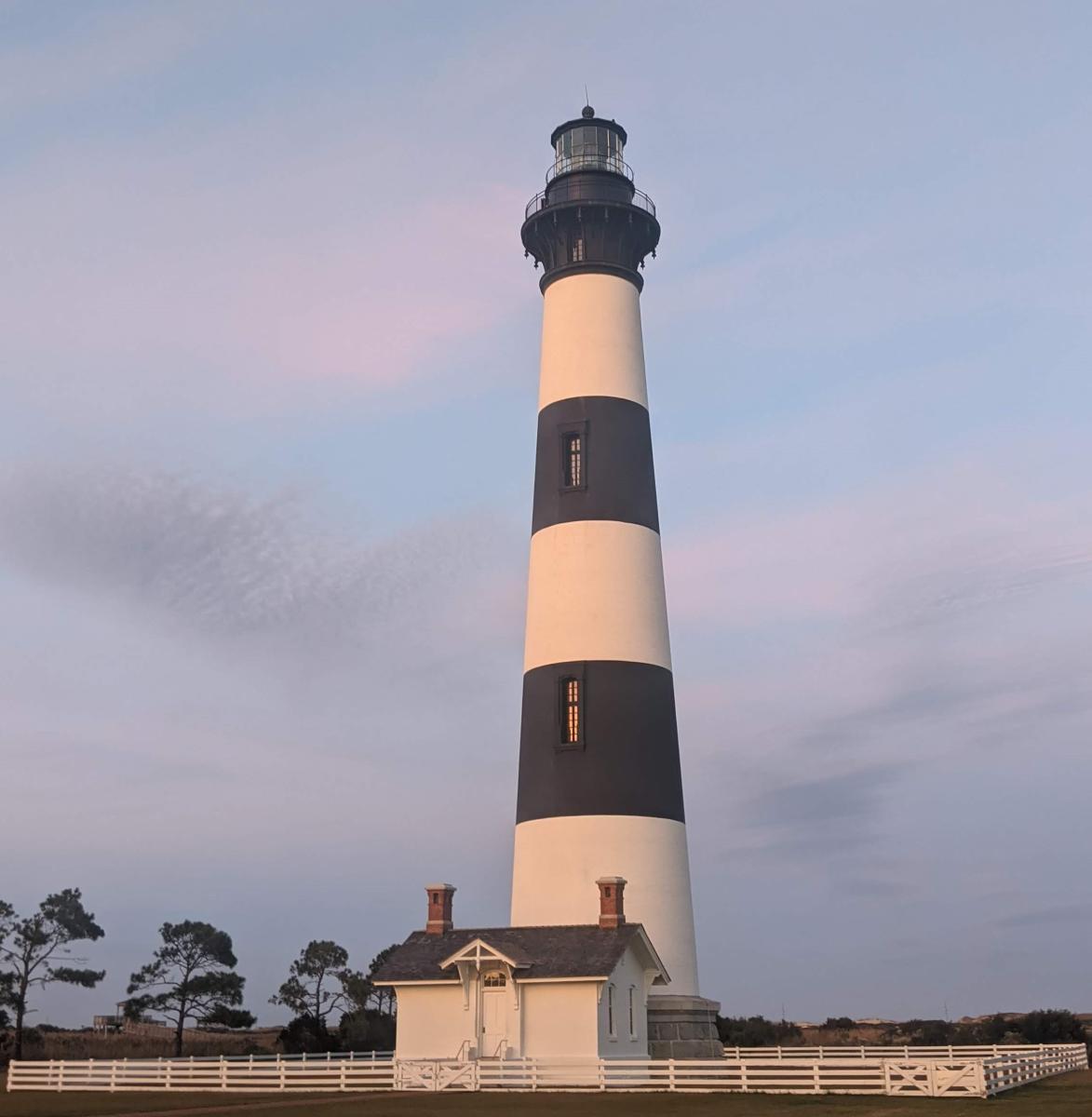 A lighthouse with a striped pattern against an evening sky. Trees and a lightkeeper's house are at the bottom of the lighthouse. 