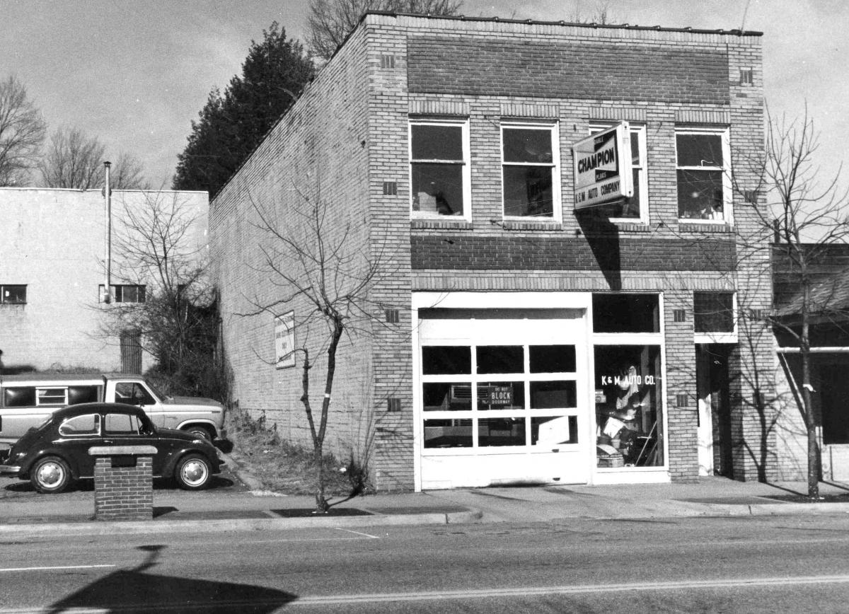 Black and white photograph of a two-story brick building with a white garage door and large window on the ground floor. 