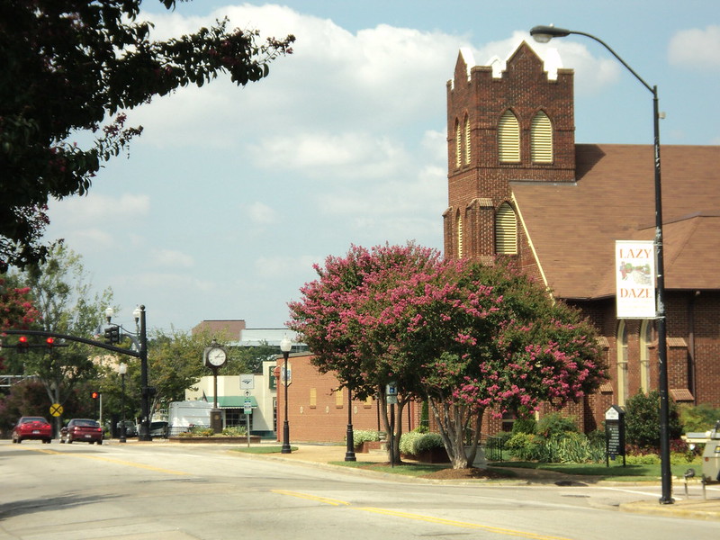 An avenue in a small downtown. A church presides over blooming trees. There is a stoplight and the sky is cloudy but blue.