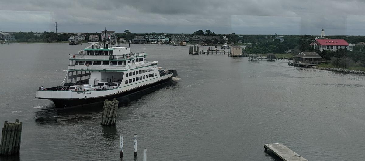 Transportation ferry crossing a waterway on a gloomy day. Wooden posts and a dock are near the ferry.