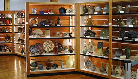 Múltiples vitrinas con cinco estantes que exhiben obras de cerámica. Las obras incluyen platos, cuencos, jarrones y cántaros.