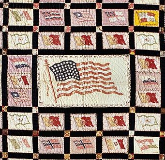 A quilt depicts different flags of indigenous tribes and countries. It has strong black borders that divide each flag from each other, and a large American flag sits in the middle of the quilt design.