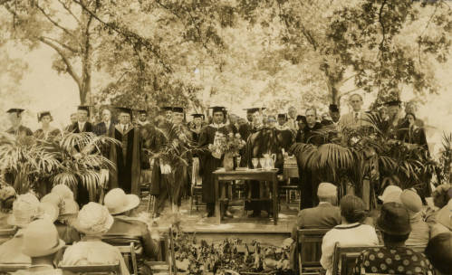 Black and white photograph of approximately 30 people wearing caps and gowns on a stage in front of an audience. Dr. David Dallas Jones stands in the middle holding a piece of paper and looking at the camera with a serious look on his face.