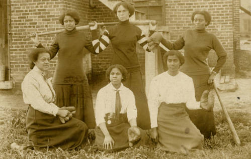 Black and white photograph of six young women holding baseball equipment. They are all wearing long skirts and long sleeved-shirts and appear to be Black women. 
