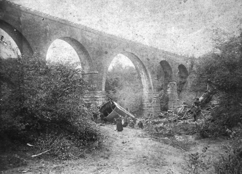 A wrecked train rests at the foot of a viaduct. Black and white photo.