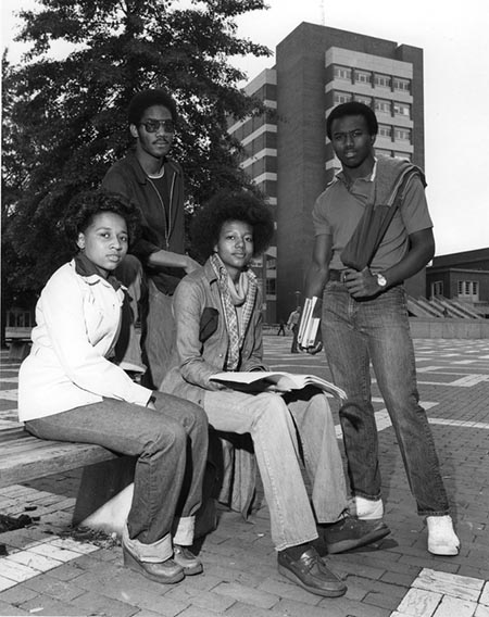 Four students sit on a bench outside a large library at a university.