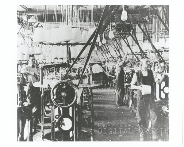 Photograph inside of Hanes Knitting Factory; workers stand by machinery.