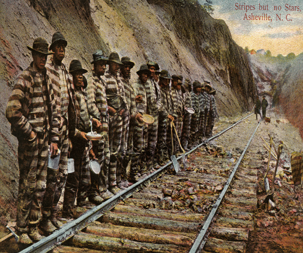 Color photograph of a row of African American men wearing white and black striped prison uniforms standing in a line alongside a railroad track. Mountains are in the background.