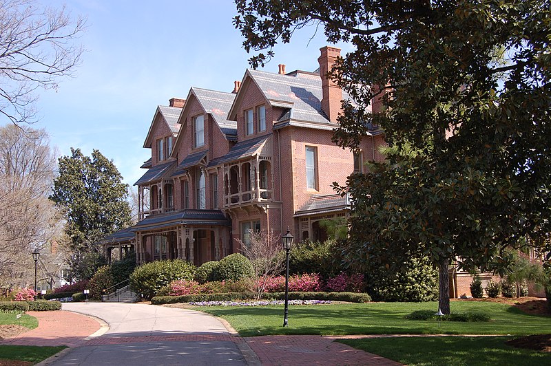 A brick mansion with three stories. It is surrounded by greenery and has a path in front.