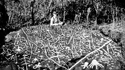 A man sits behind Judaculla rock, a boulder engraved with images that are here highlighted with chalk or paint. This boulder was considered sacred by native tribes from the time of the earliest settlers' arrival. Judaculla rock is located outside of present-day Sylva. This image was taken between 1945 and 1949. Courtesy of the North Carolina Museum of History.  