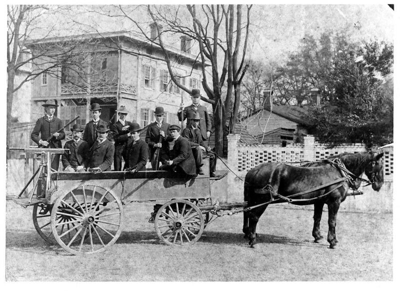 Ten armed men sit in a carriage drawn by a horse with a machine gun mounted on the back. A house is in the background. Black and white photo.
