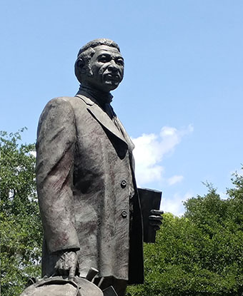 Stone statue depicting Denmark Vesey holding his hat and book on a sunny day surrounded by trees
