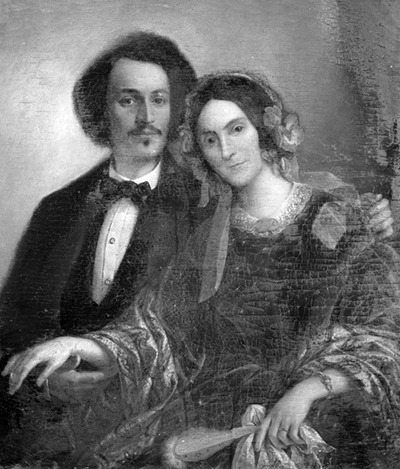 John and Peggy in portrait. John has longer hair, and moustache-goatee is wearing a suit. Peggy is wearing a hair piece with a dress and has medium-length hair.