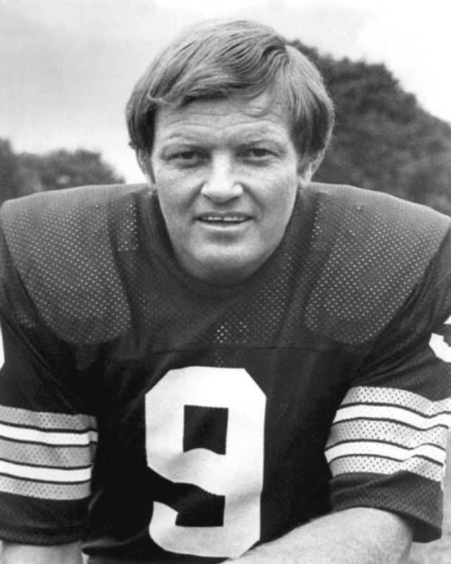 Black and white photograph of a white man with medium-length hair in a dark-colored football jersey. The man, Sonny Jurgensen, is kneeling and the photograph is taken from the waist up.