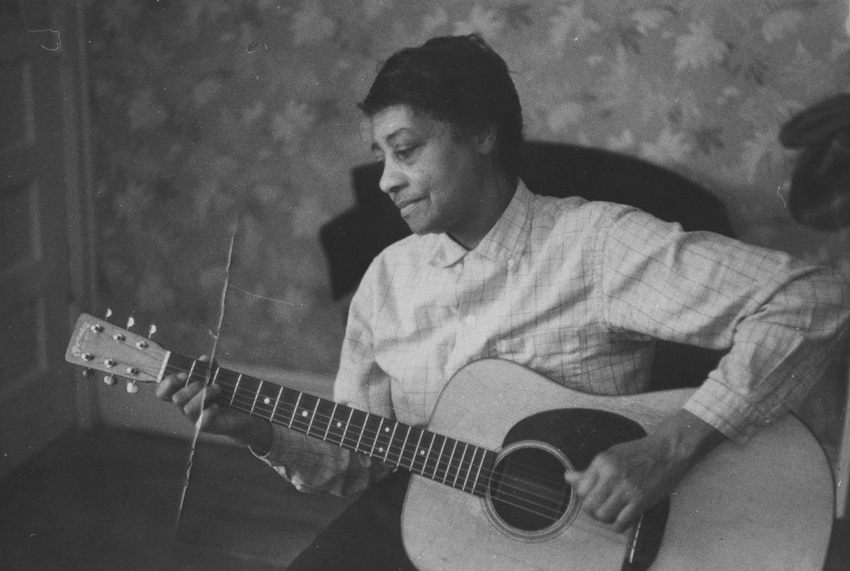Black and white photo of an older black woman wearing a loose braid and a flannel button-up shirt. She seated and holding a guitar in her lap with her left hand on the body of the guitar and her right hand on the frets.