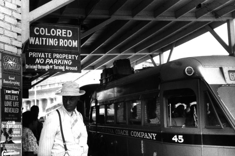 A black and white photo of a black man standing in front of a sign that reads "Colored Waiting Room" in front of a train. 