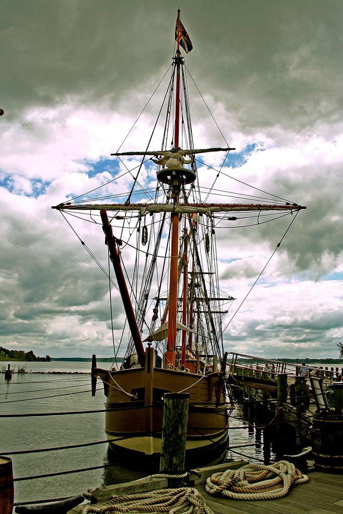 Sailing ship in Jamestown. It is docked in a harbor. It is clad with several masts and ropes for the masts. It is a cloudy day.