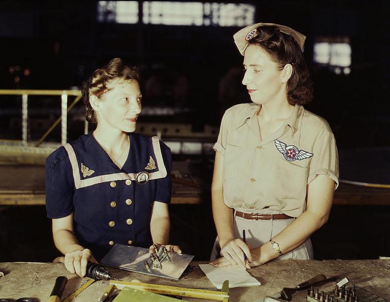 Two women stand beside each other on a factory floor. They are wearing military-like apparel.