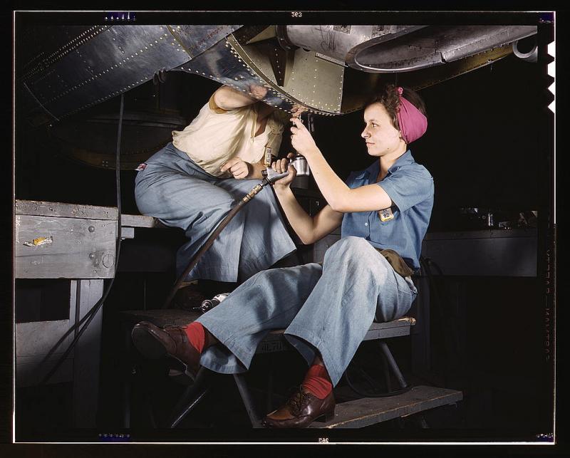 Two women in worksuits construct a bomber airplane. Their hair is tied up and they have tools in their hands.