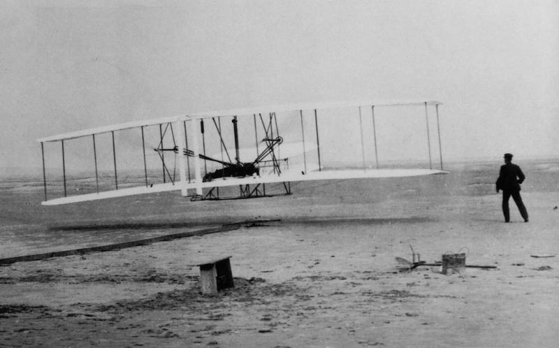 A rudimentary airplane hovers over sand. Men stand alongside to try to operate it. 