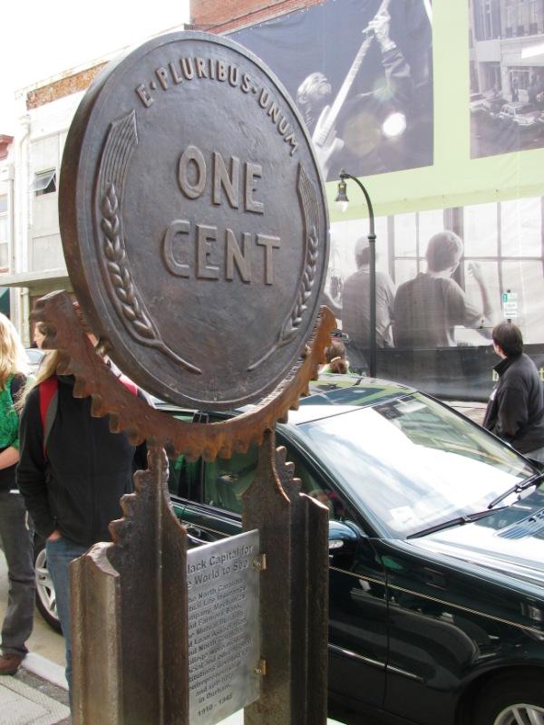 Large one cent bronze marker. It is on a pedestal adjacent to the street. There are people and cars around it. It is sunny. 