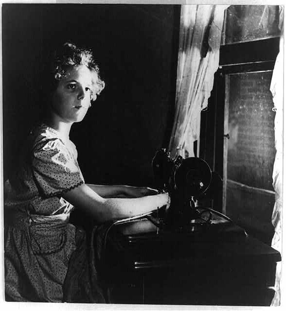 A young girl sits beside a window and an electric sewing machine. She is wearing a dress. Black and white photo.