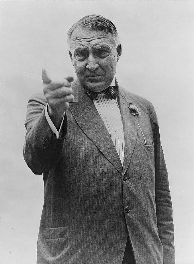 Warren G. Harding. Black and white. He is wearing a suit and pointing his finger at the camera. He has a furrowed brow and short hair. He has no facial hair. 
