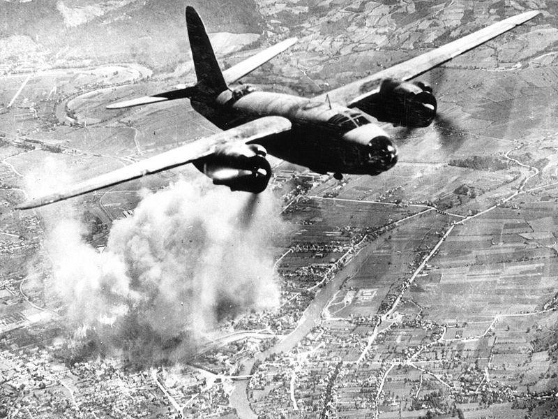 Black and white photo of a bomber flying over land. 