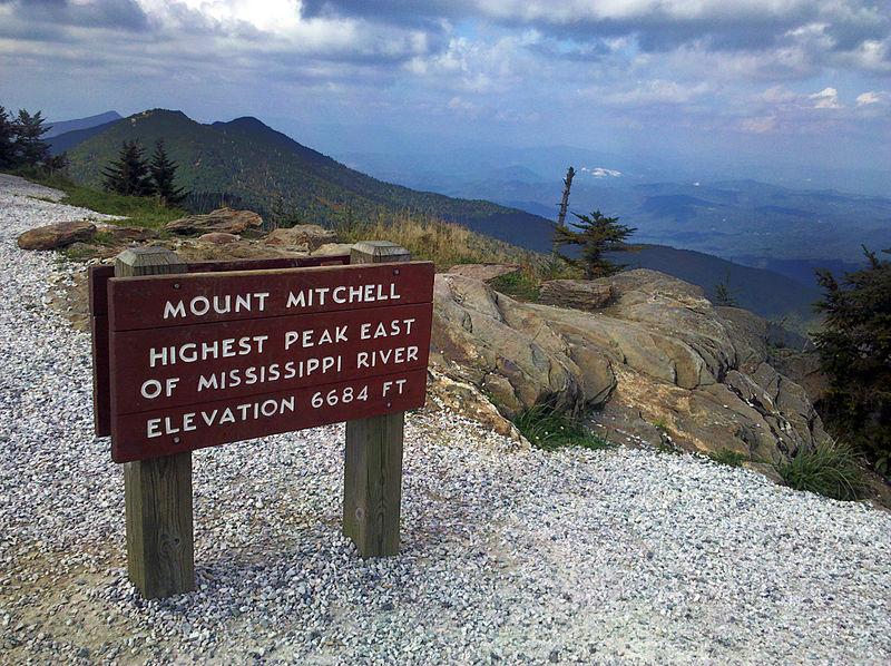 Mount Mitchell peak with Mount Craig behind. The sign is red and there are clouds in the sky. The mountaintops are green. 