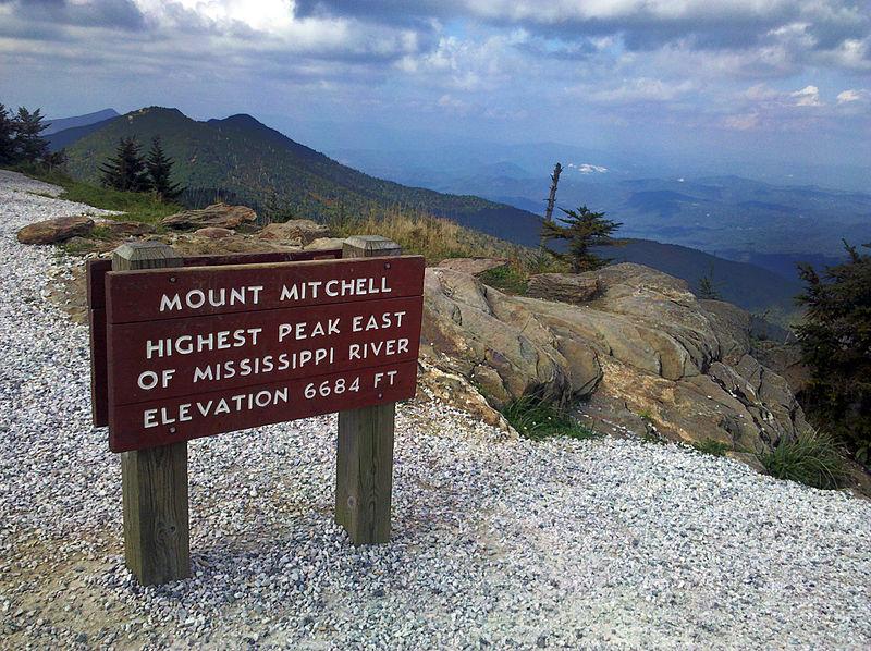 Mount Mitchell Mount Mitchell peak with Mount Craig behind. The sign is red and there are clouds in the sky. The mountaintops are green.