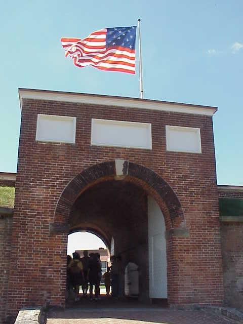 Fort McHenry entrance. It is a brick archway with walls attached. An American flag flies overhead against a clear sky. 