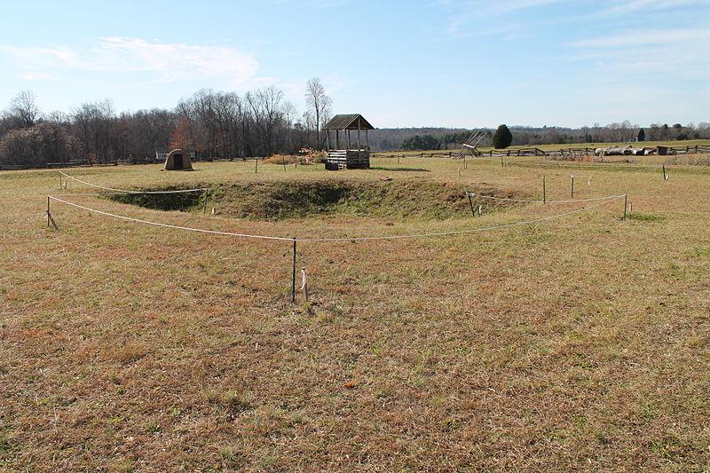 An open field with depressed craters. A small wooden well is in the background and a treeline marks the edge of the landscape.